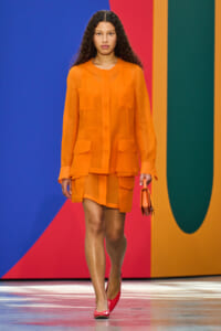 Model walking the runway in an orange sheer blouse and matching shorts, holding a small orange bag against a colorful geometric backdrop.