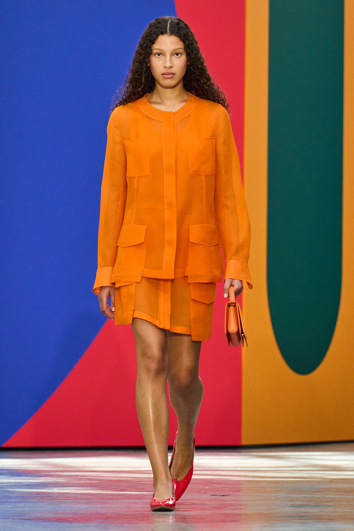 Model walking the runway in an orange sheer blouse and matching shorts, holding a small orange bag against a colorful geometric backdrop.