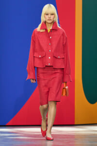 Runway model wearing a red cropped jacket and matching shorts in a bold red hue, yellow top visible at the collar, red heels, and orange handbag, against a colorful backdrop.