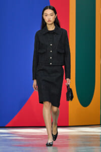 Female model walks a runway in a black denim jacket with silver buttons and a matching knee-length skirt, holding a small black handbag against a bold geometric backdrop.
