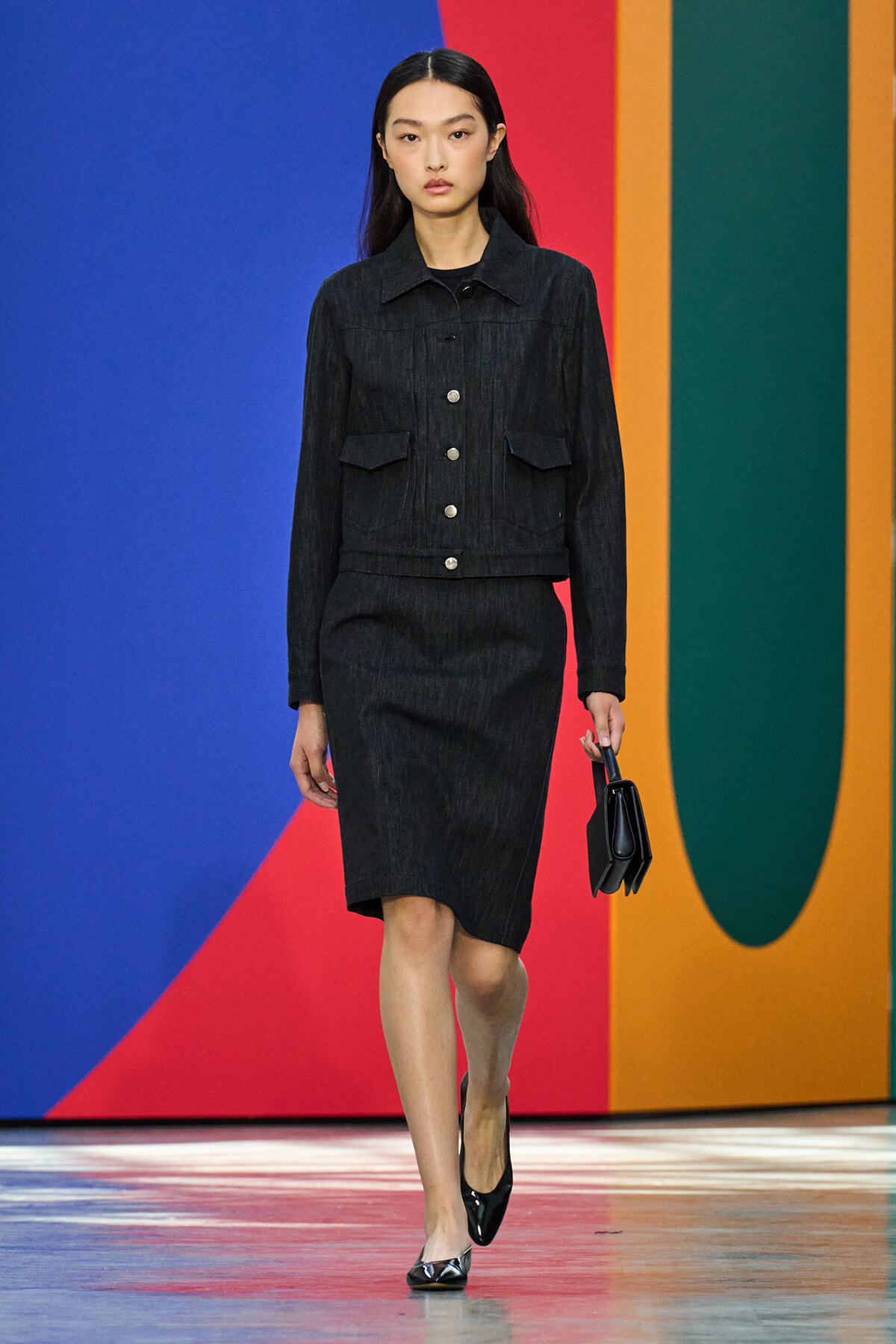 Female model walks a runway in a black denim jacket with silver buttons and a matching knee-length skirt, holding a small black handbag against a bold geometric backdrop.
