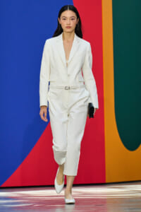 Model walking the runway in a white tailored suit with belt and white heels, holding a black clutch, against a bright, colorful background.