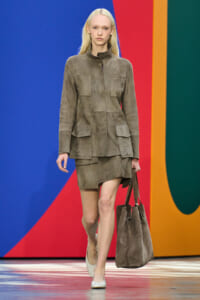 Model walks a runway wearing a taupe suede utility jacket with multiple pockets and a matching mini skirt, carrying a large taupe suede tote.