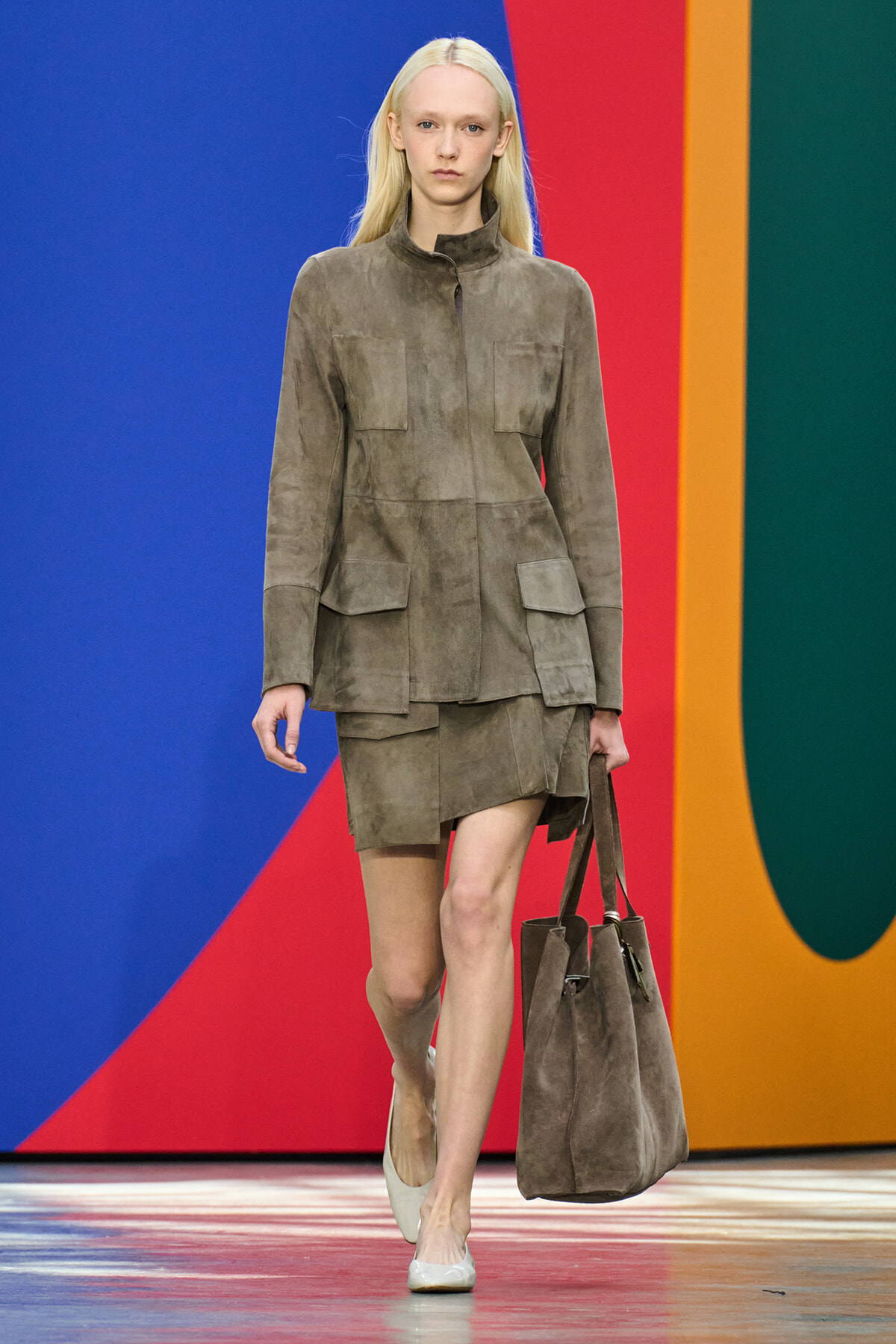Model walks a runway wearing a taupe suede utility jacket with multiple pockets and a matching mini skirt, carrying a large taupe suede tote.