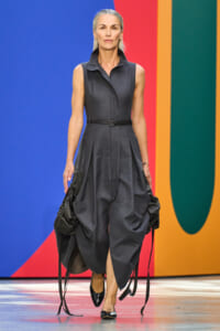 Female model walking on a fashion runway in a sleeveless dark gray belted dress with a high collar and sculpted pleats, against a bright colorful backdrop.
