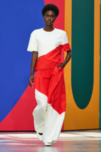 Model walking the runway in a white and red color-block top and wide-leg pants against a bold geometric backdrop.