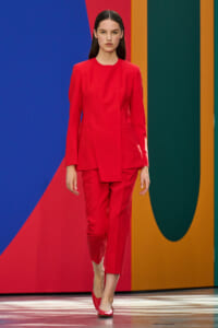 Female model in a bright red tailored suit walking the runway against a colorful geometric backdrop.