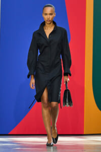 Model in a black, long-sleeve dress with a front slit walks the runway against a colorful abstract backdrop, carrying a black handbag and wearing black heels.