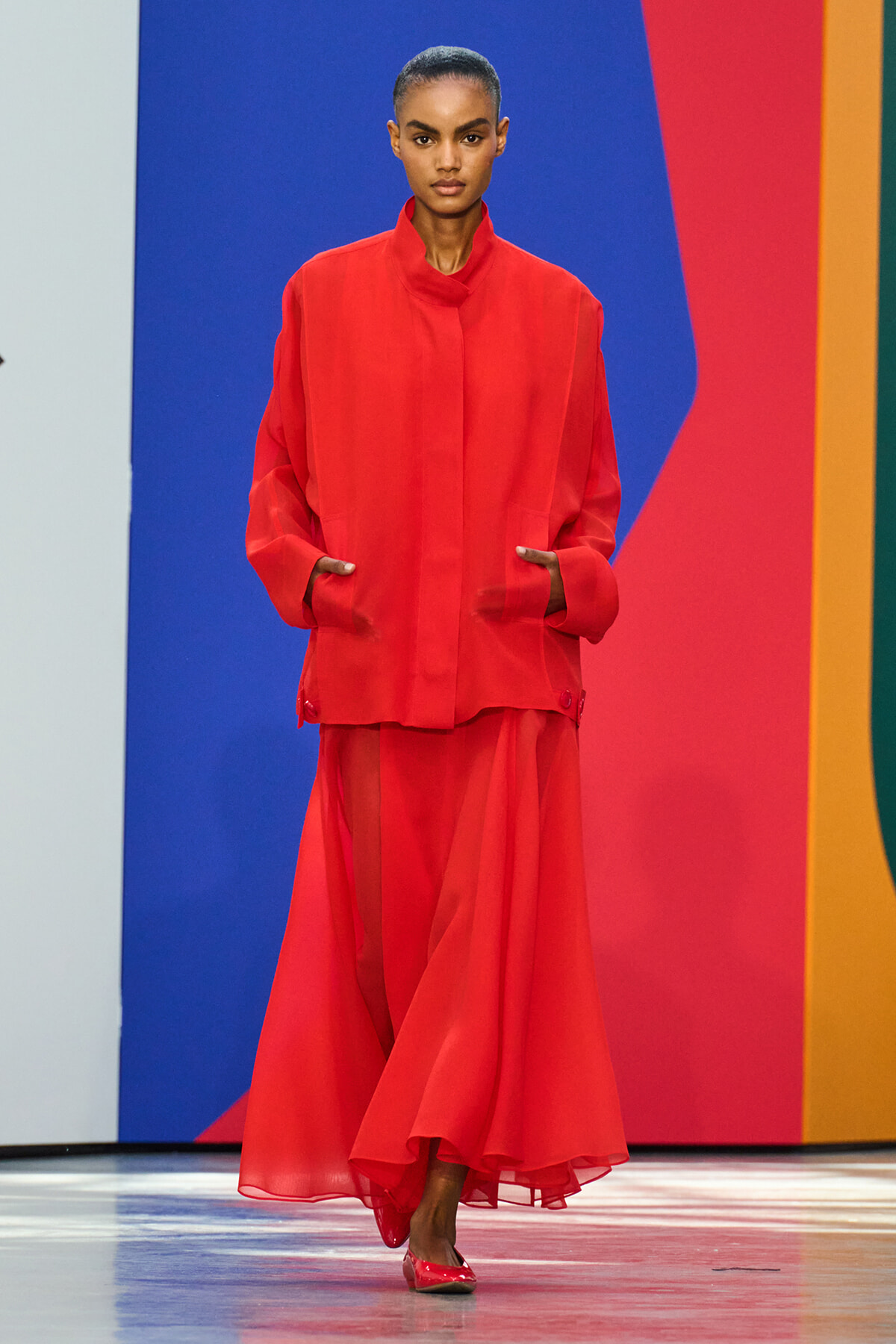 Model in a bold red ensemble—a loose red coat over a flowing red dress—walking a runway with a colorful backdrop of blue, red, and orange Panels.