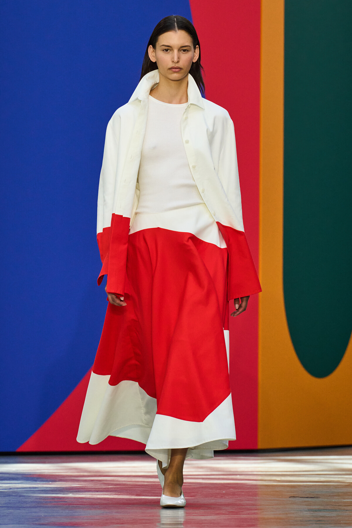 Model walking a fashion runway in a color-block white and red gown with a white jacket, blue and multicolored backdrop behind her