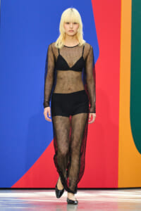 Model walking a fashion runway in a sheer black fishnet dress over a black bra and shorts, with platinum blonde hair, against a bright geometric backdrop.