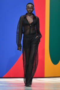 Model walks a fashion runway in a sheer black mesh outfit; bold color-block backdrop in blue, red, orange, and green.