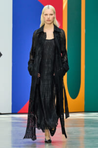 Model wearing a black striped fringe coat over a matching dress on a fashion runway with a bright geometric backdrop.