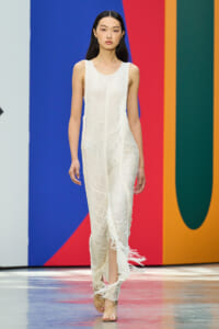 Model on a runway wearing a sleeveless white textured fringe gown, walking against a bold blue, red, and orange backdrop.
