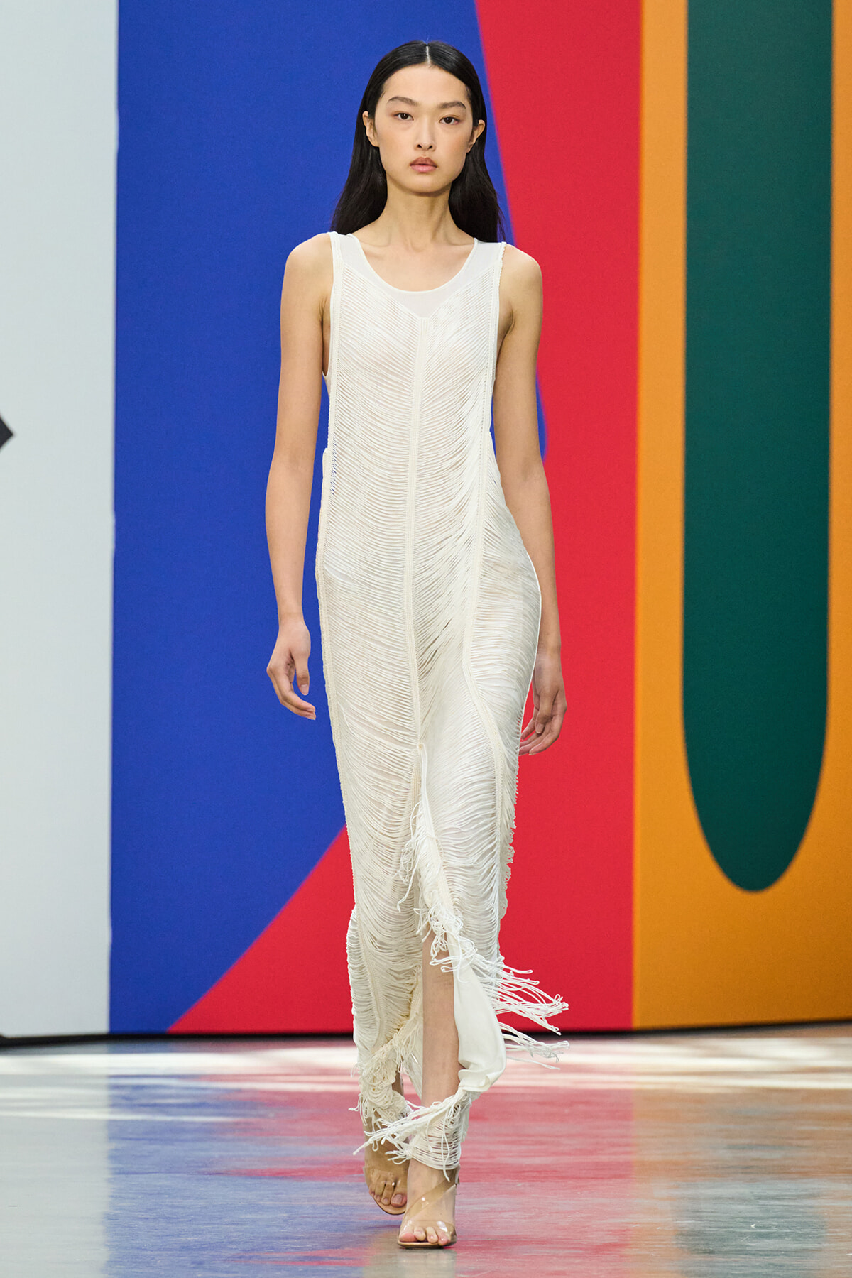 Model on a runway wearing a sleeveless white textured fringe gown, walking against a bold blue, red, and orange backdrop.