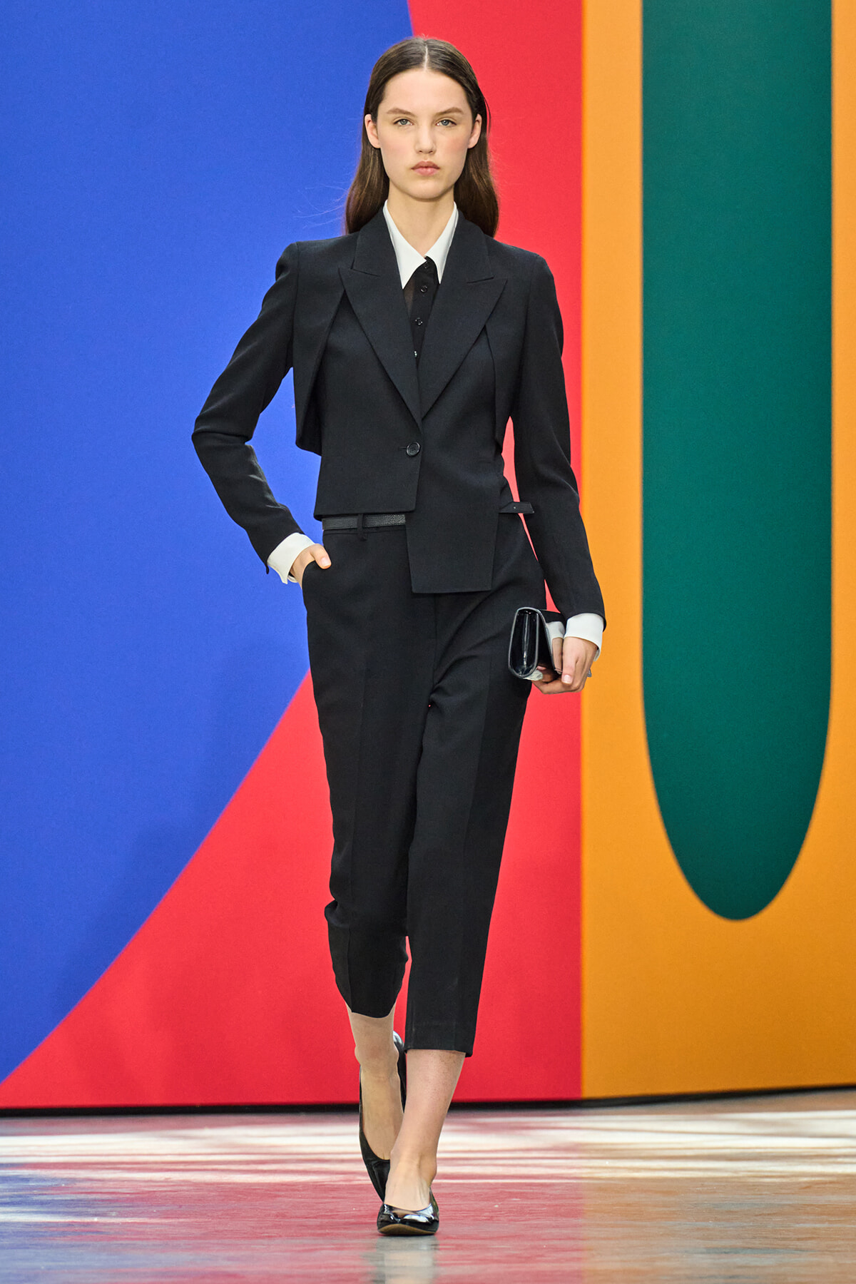 Model walking the runway in a black tailored pantsuit with white shirt and black tie, holding a small black clutch against a bold color-block backdrop.