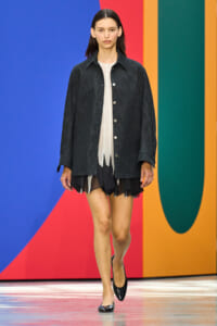 Model walking runway in dark denim jacket over a white top and black layered skirt; colorful geometric backdrop behind her.