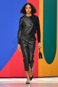 Female model walks a fashion runway in a black leather color-block outfit, holding a small black handbag, against a bright geometric backdrop.
