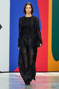 Model walking a fashion runway in a black sheer striped cardigan and matching pants against a colorful backdrop.