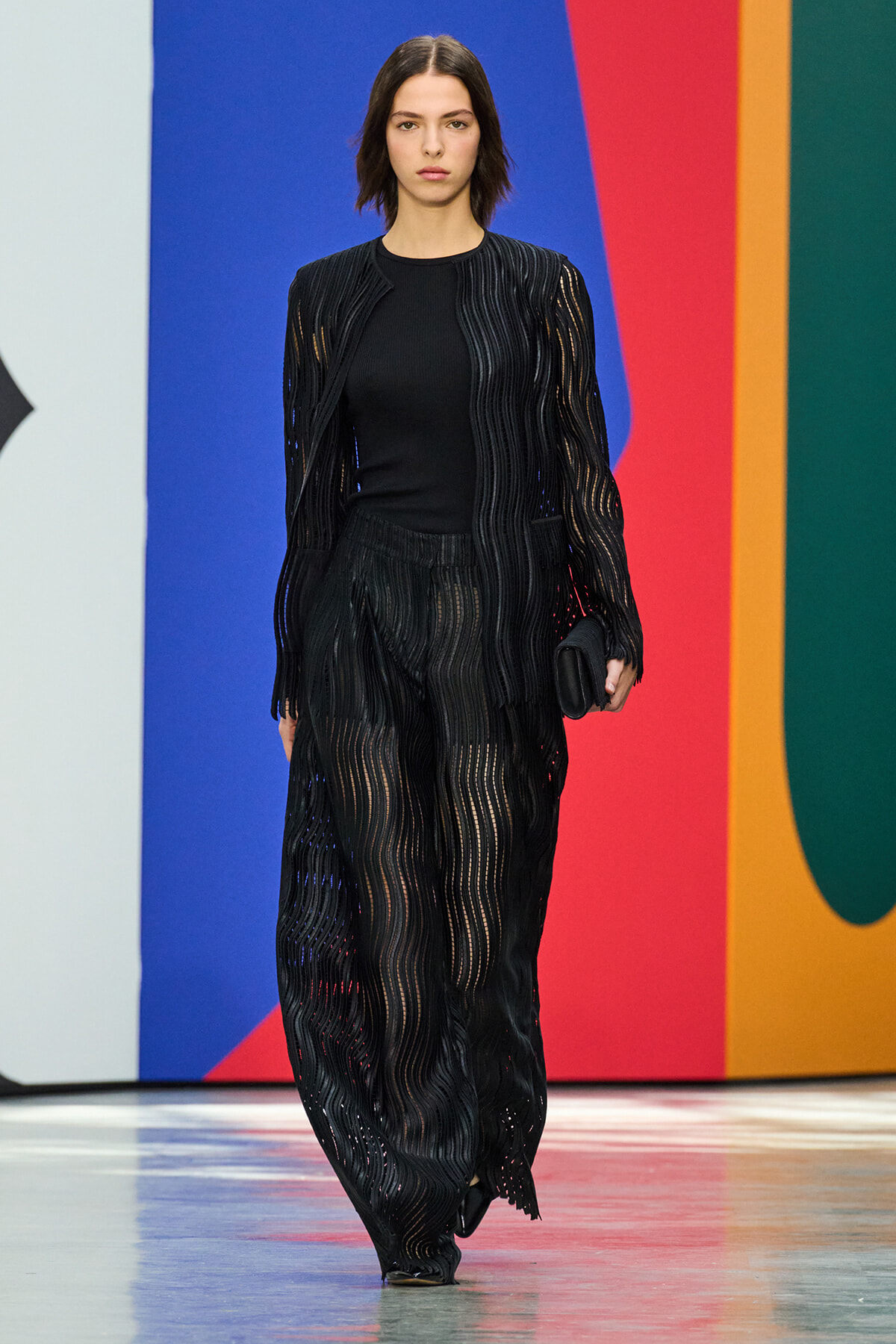 Model walking a fashion runway in a black sheer striped cardigan and matching pants against a colorful backdrop.