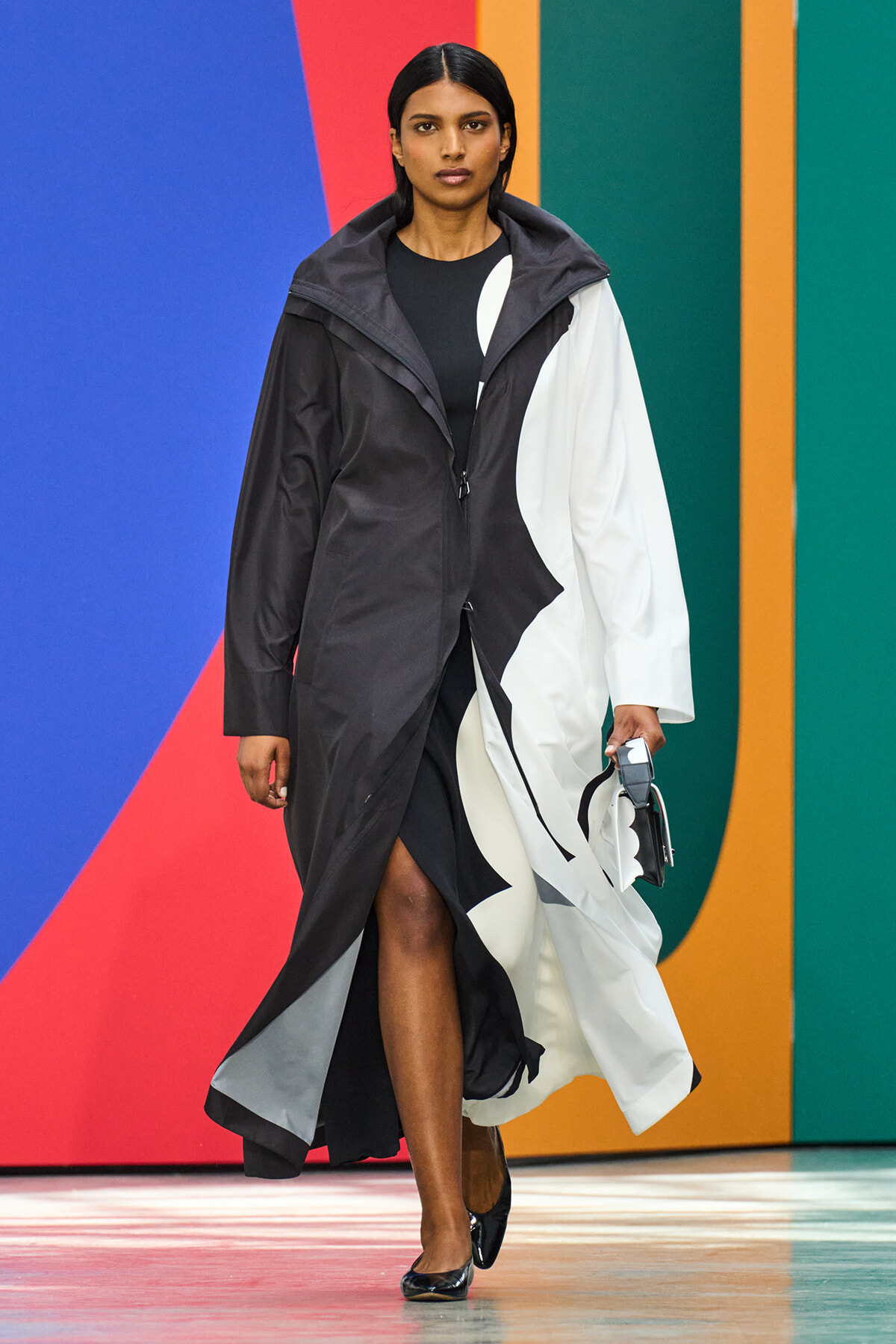Model walking the runway in a long black-and-white color-block coat over a black dress, holding a small handbag, against a vibrant geometric backdrop.