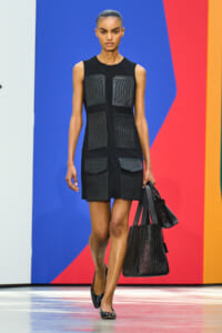 Model walking runway in a black sleeveless dress with geometric textured panels, carrying matching black bags, against a colorful background