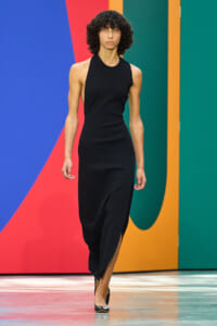 Model walking the runway in a sleeveless black midi dress against a bold multicolored geometric backdrop.