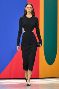 Model in a black long-sleeve dress with side cutouts walking on a colorful runway backdrop holding a small clutch.
