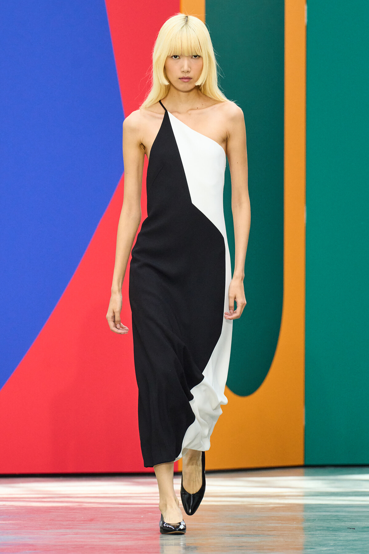 Model walks the runway in a black-and-white color-block dress against a bright geometric backdrop.