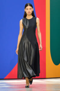 Model wearing a sleeveless black and dark leather color-block dress walks the runway against a bold blue, red, orange, and green backdrop.