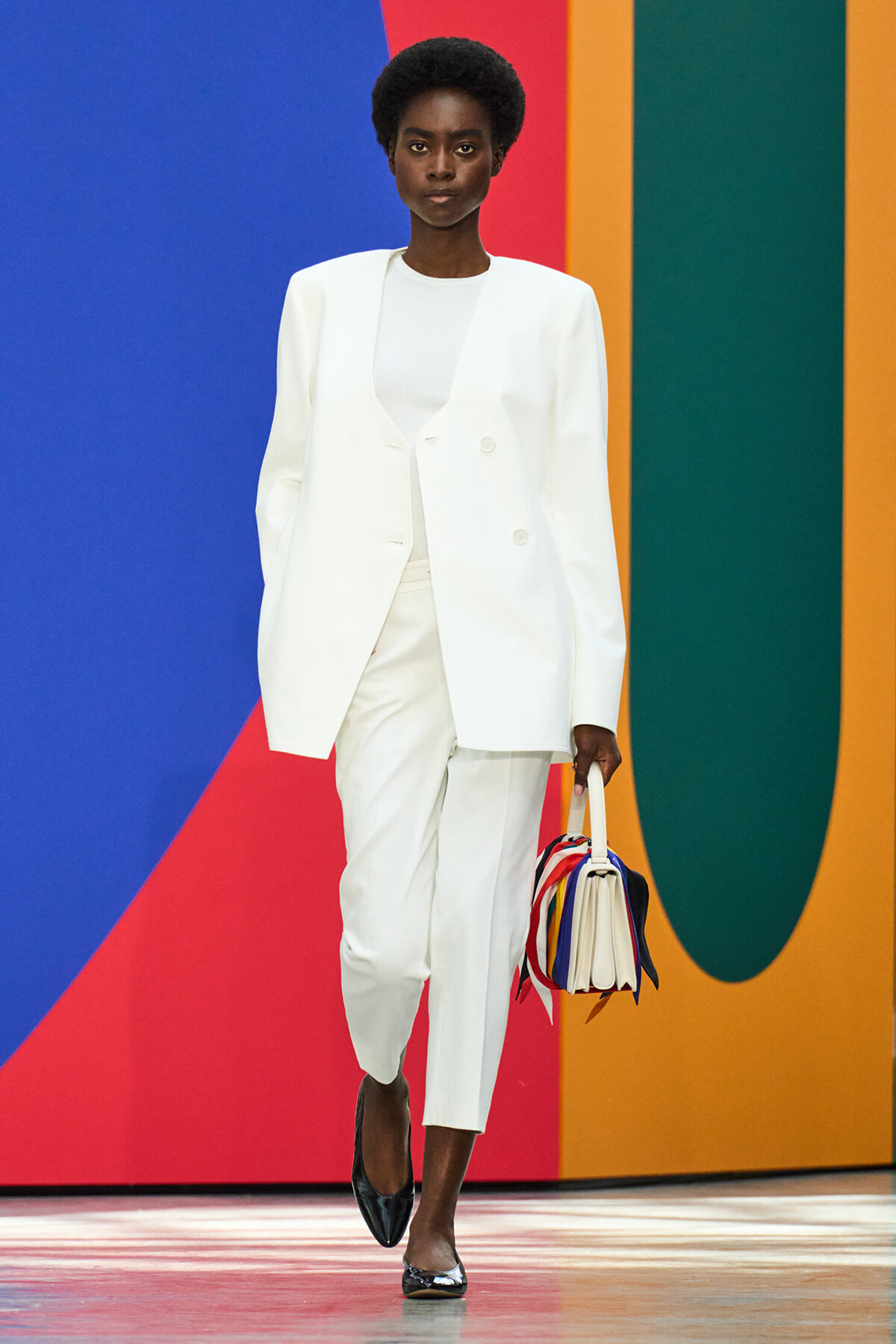 Woman model wearing a white tailored pantsuit and white top, walking with a multicolored handbag against a bold blue, red, orange, and green backdrop