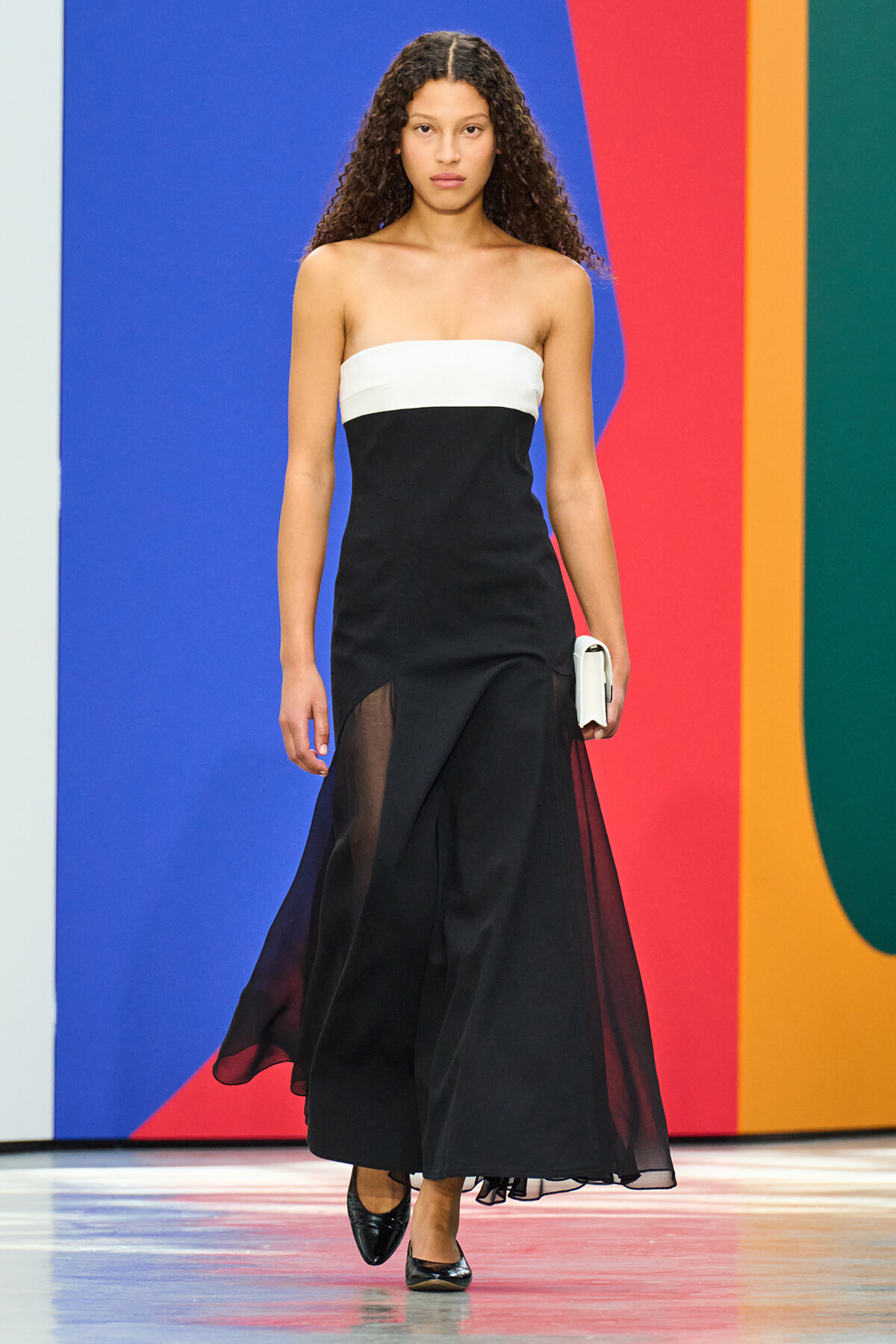 Model walking on a runway wearing a strapless black dress with a white top band and sheer panels, holding a white clutch.