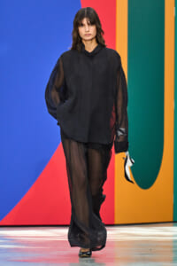 Model walks a black sheer oversized blouse and wide-leg pants on a fashion runway with a bold, colorful striped backdrop.