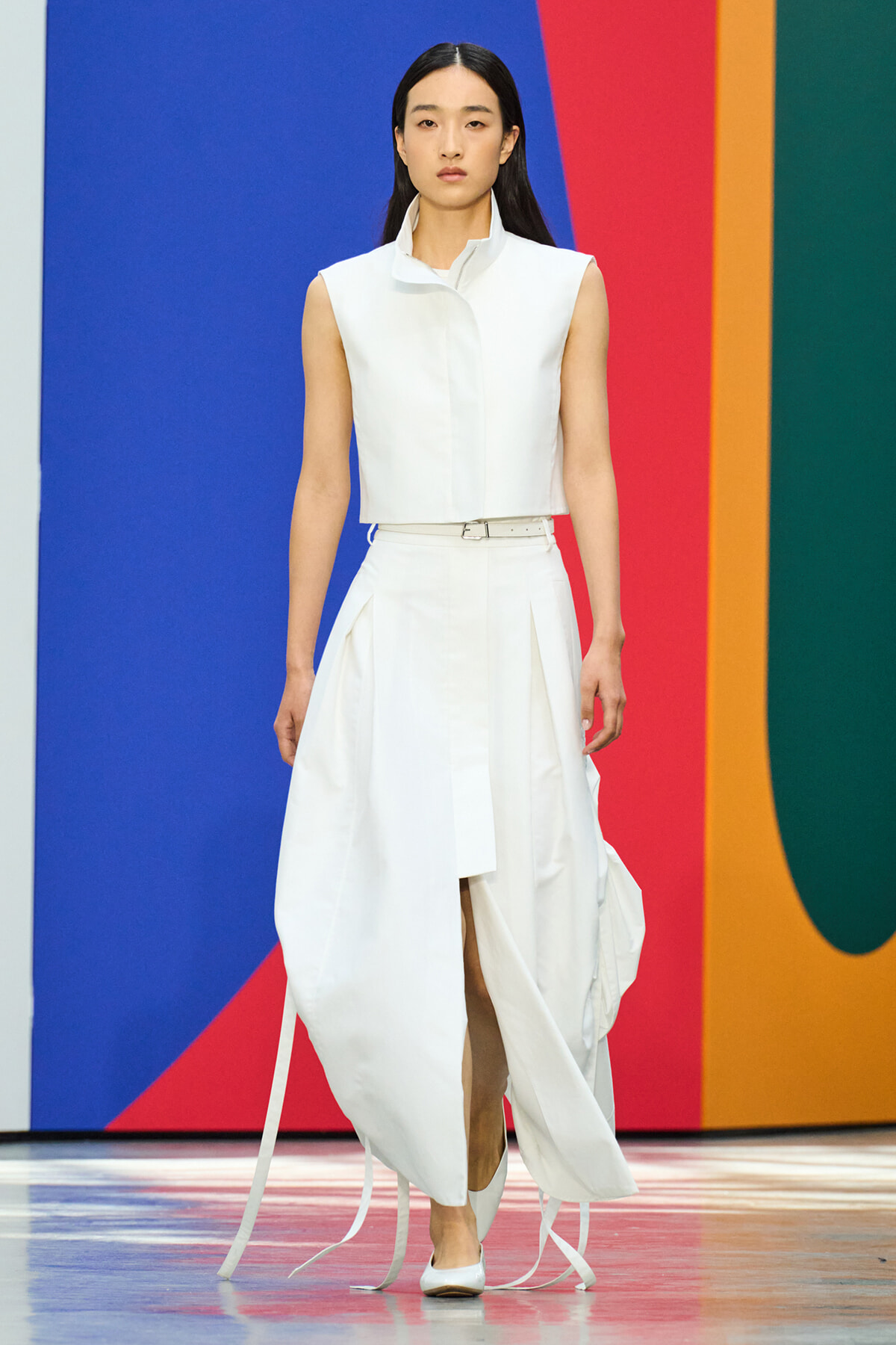Model on a runway wearing a white sleeveless cropped top and high-waisted, draped long skirt with a belt, against a bold multicolor backdrop.
