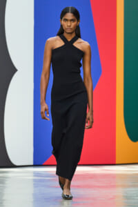 Model walking a runway in a black halter-neck dress against a colorful abstract backdrop, during a fashion show.