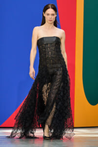 Model walking runway in a strapless black gown with a geometric mesh overlay against a colorful backdrop