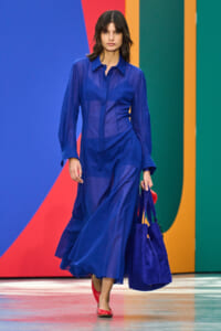Runway model in a cobalt blue sheer blouse and matching flowing skirt, carrying a blue handbag against a bright, multicolored backdrop.