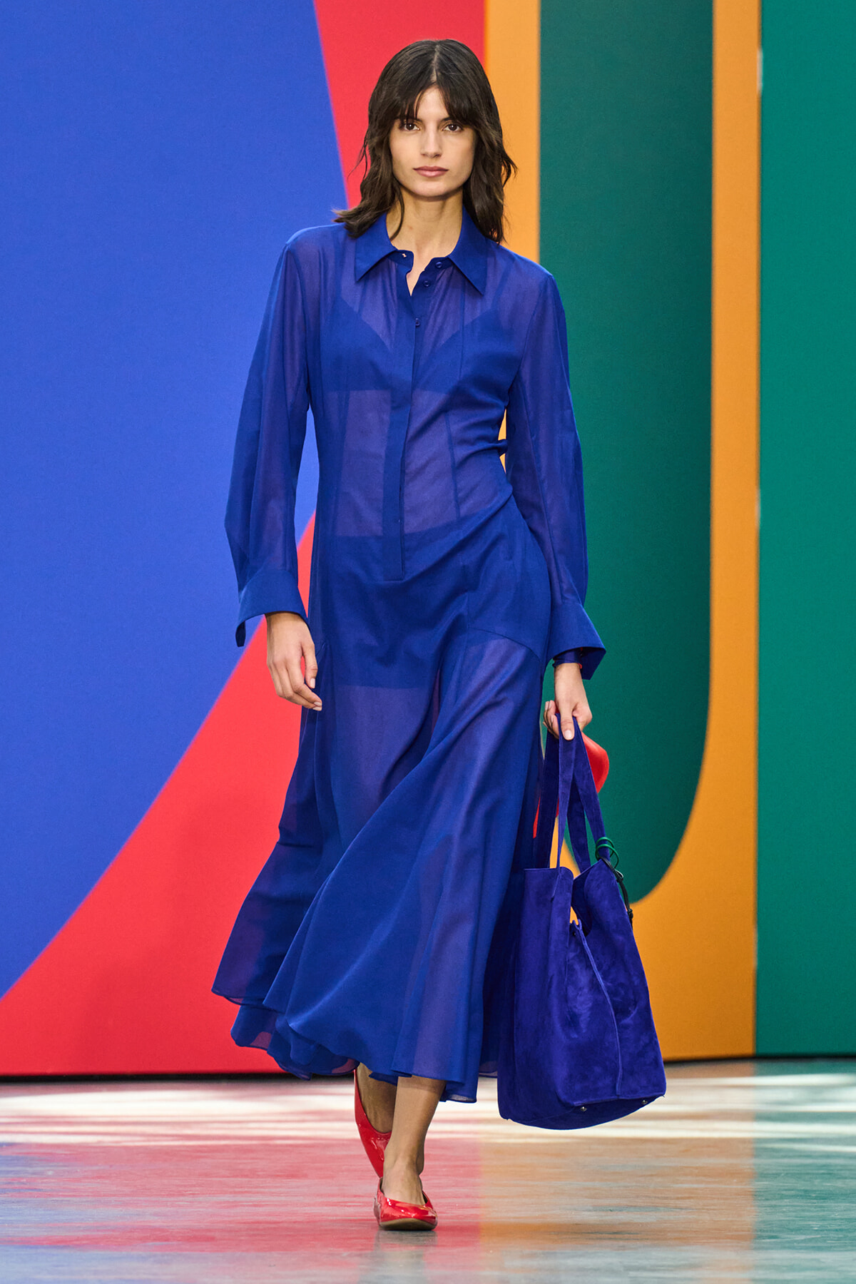 Runway model in a cobalt blue sheer blouse and matching flowing skirt, carrying a blue handbag against a bright, multicolored backdrop.
