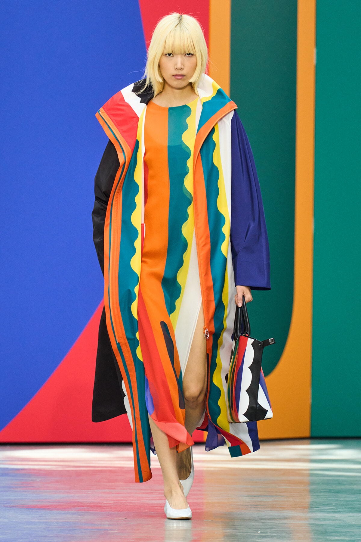 Model walking a runway in a long, multicolor striped coat with wavy yellow accents, holding a matching striped bag.