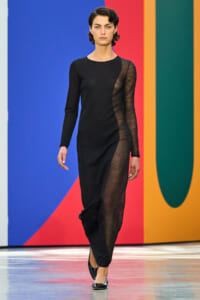 Female model walking a runway in a black long-sleeve dress with sheer mesh panels against a colorful striped backdrop.