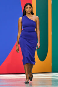 Woman in a royal blue one-shoulder midi dress walks the runway against a bold, multicolored geometric backdrop.