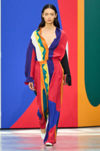 Female model on a fashion runway wearing a bold color-block jumpsuit in red, blue, orange, yellow, green; she holds a small clutch.