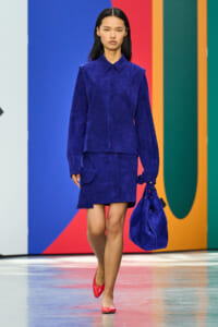 Model walks a runway in a cobalt blue suede two-piece outfit, carrying a matching blue bag and wearing red flats against a colorful backdrop.