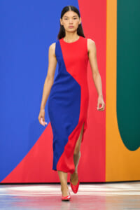Female model walks down a runway in a red-and-blue color-block sleeveless midi dress with a diagonal panel, paired with red heels, against a multicolor backdrop.