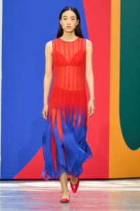 Model walking a runway in a sleeveless red sheer dress with a blue fringe hem, paired with red flats, against a bold color-block backdrop.