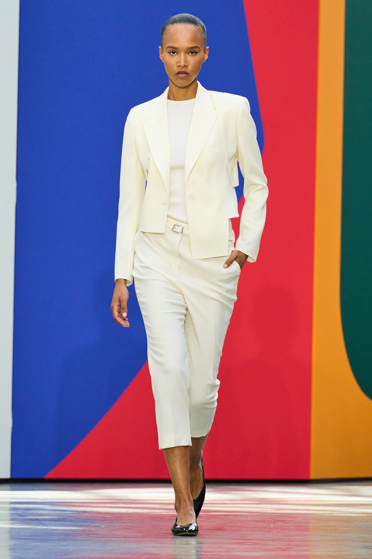 Model walking the runway in a white tailored blazer and cropped pants, with black heels, against a bright multi-color backdrop.