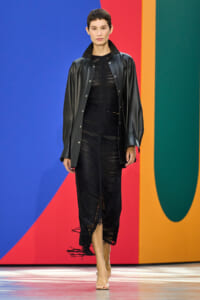 Model walking a runway in a black leather jacket over a sheer black ruched dress with fringe hems, neutral backdrop of bold color blocks behind her