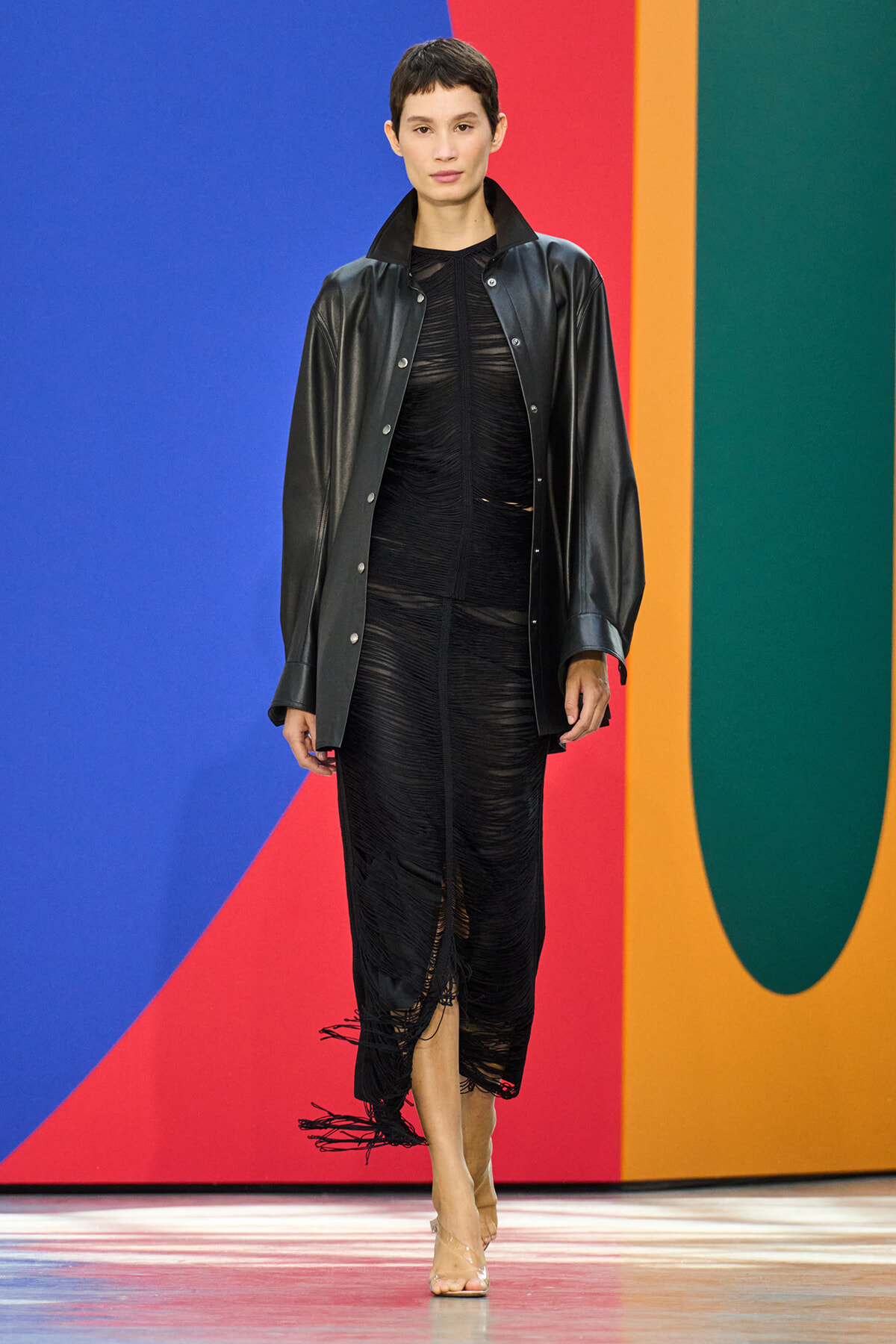 Model walking a runway in a black leather jacket over a sheer black ruched dress with fringe hems, neutral backdrop of bold color blocks behind her