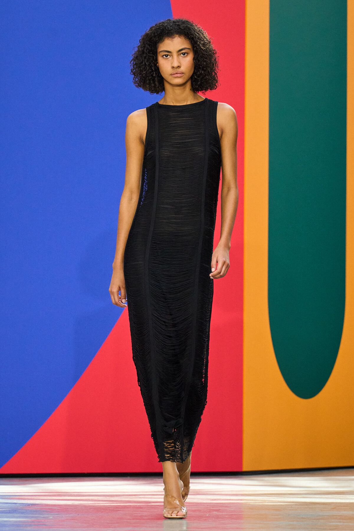Model walks a black textured sleeveless dress on a runway with a bold, colorful geometric wall in blue, red, orange, and teal behind her.