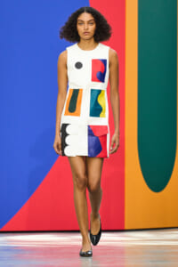 Model walks the runway wearing a white color-block dress with bold geometric shapes in red, blue, orange, and black against a vibrant backdrop.